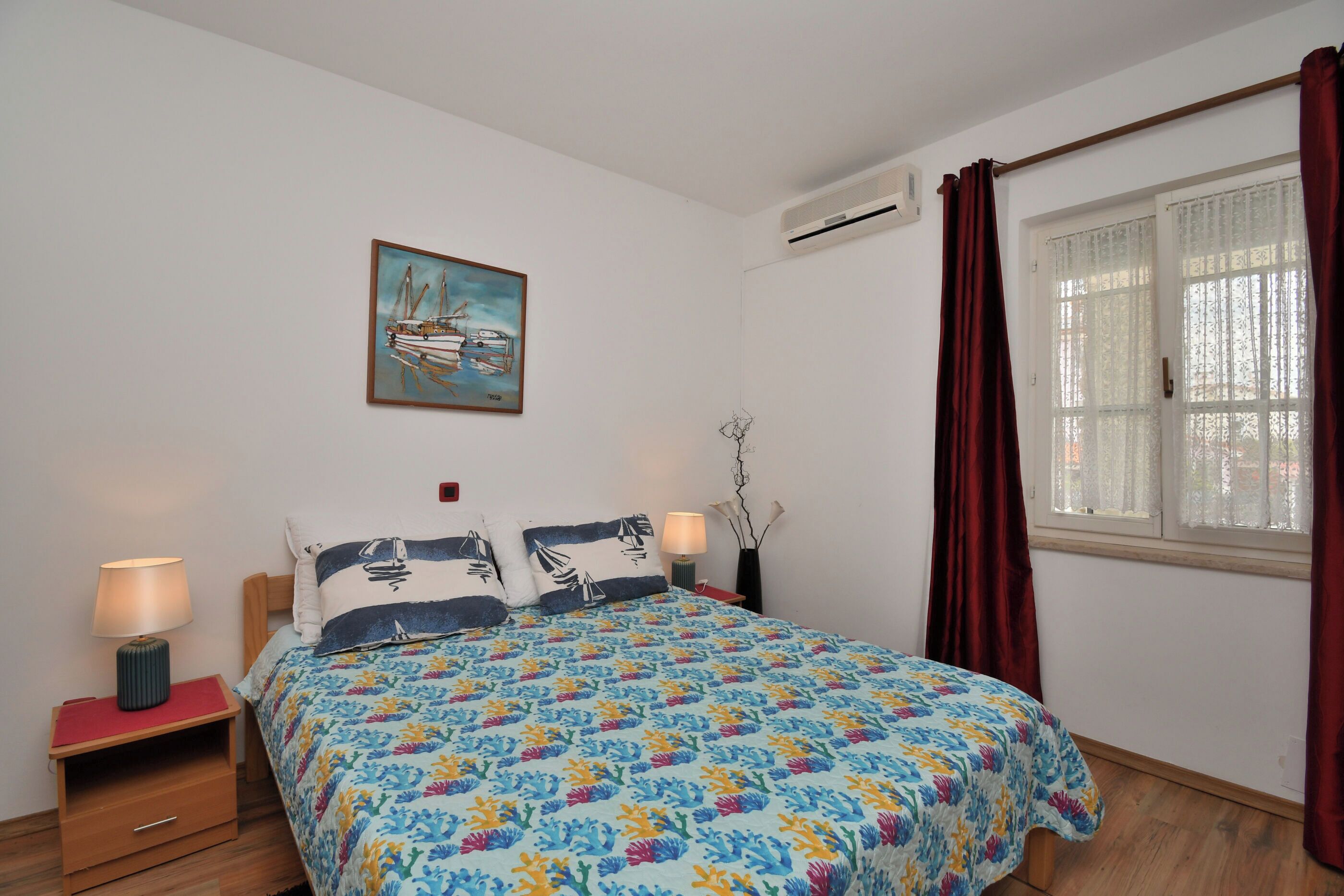 Ein Schlafzimmer mit einem Bett, zwei Nachttischen, einem Fenster mit Vorhängen und einer Wand-Klimaanlage.