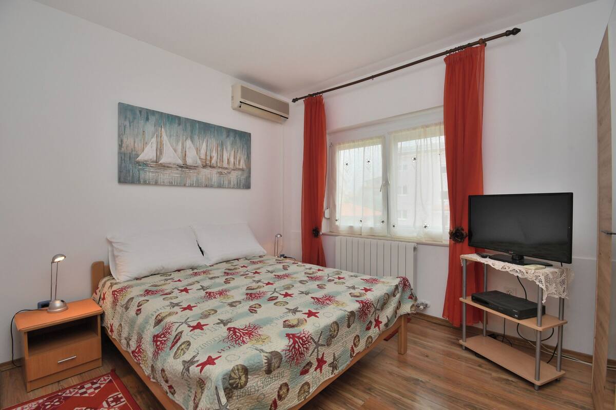 Ein Schlafzimmer mit Bett, Nachttisch, einem Fernsehgerät auf einem Schrank, einem Bild an der Wand und einem Fenster mit Vorhängen.