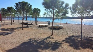 En strand med småsten, bänkar och träd, en klar himmel och ett lugnt hav.