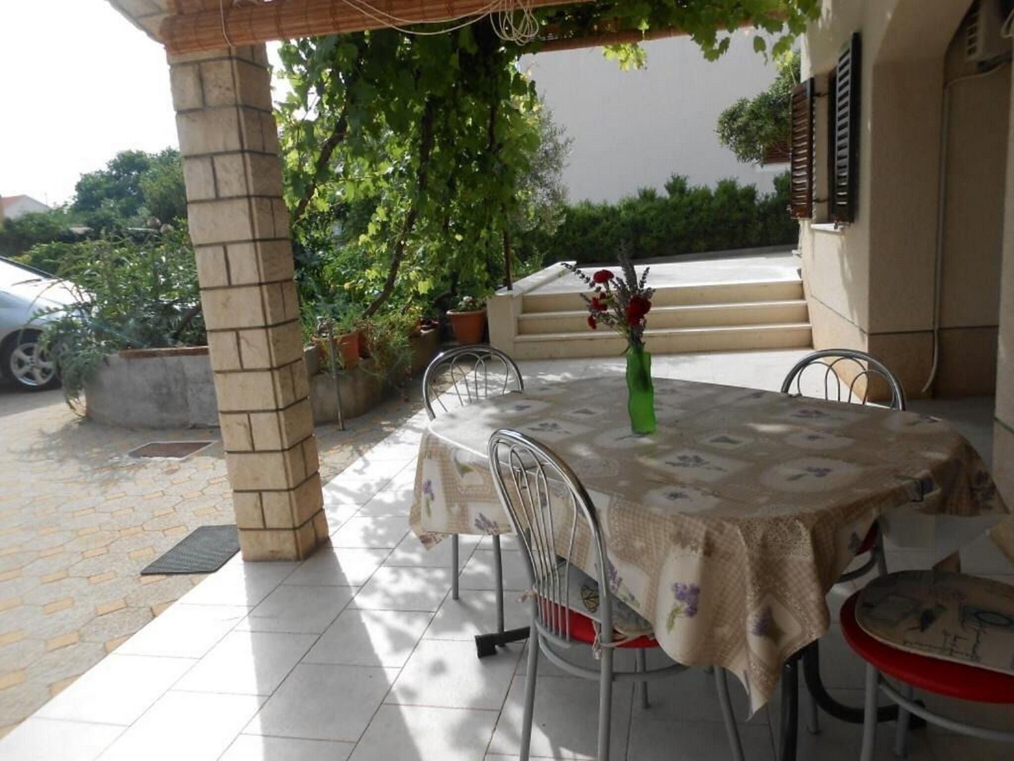 An outdoor patio with a table and chairs, surrounded by potted plants and a car in the background.