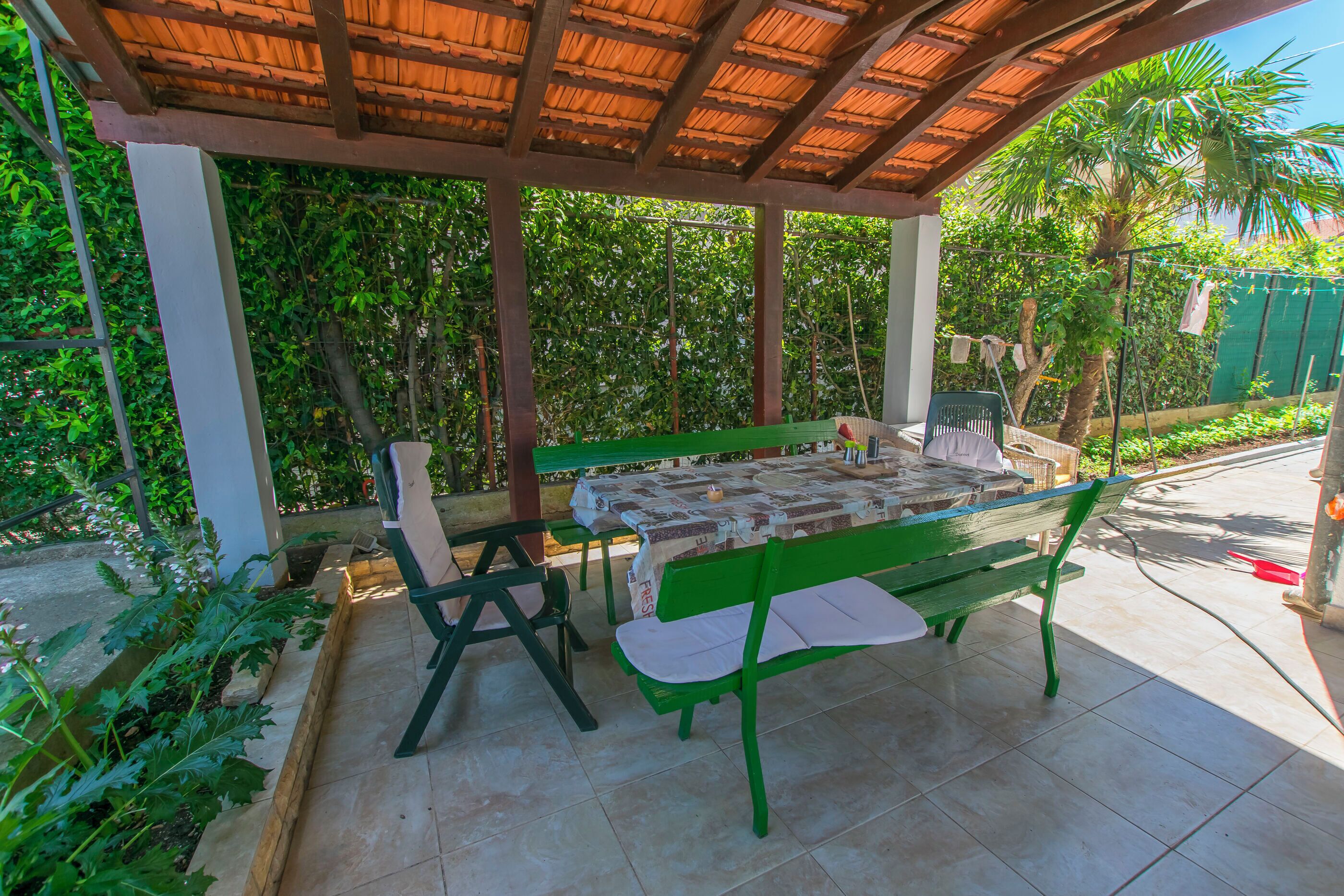 Eine überdachte Terrasse mit Tisch und Bänken, umgeben von Pflanzen und einem gefliesten Boden.