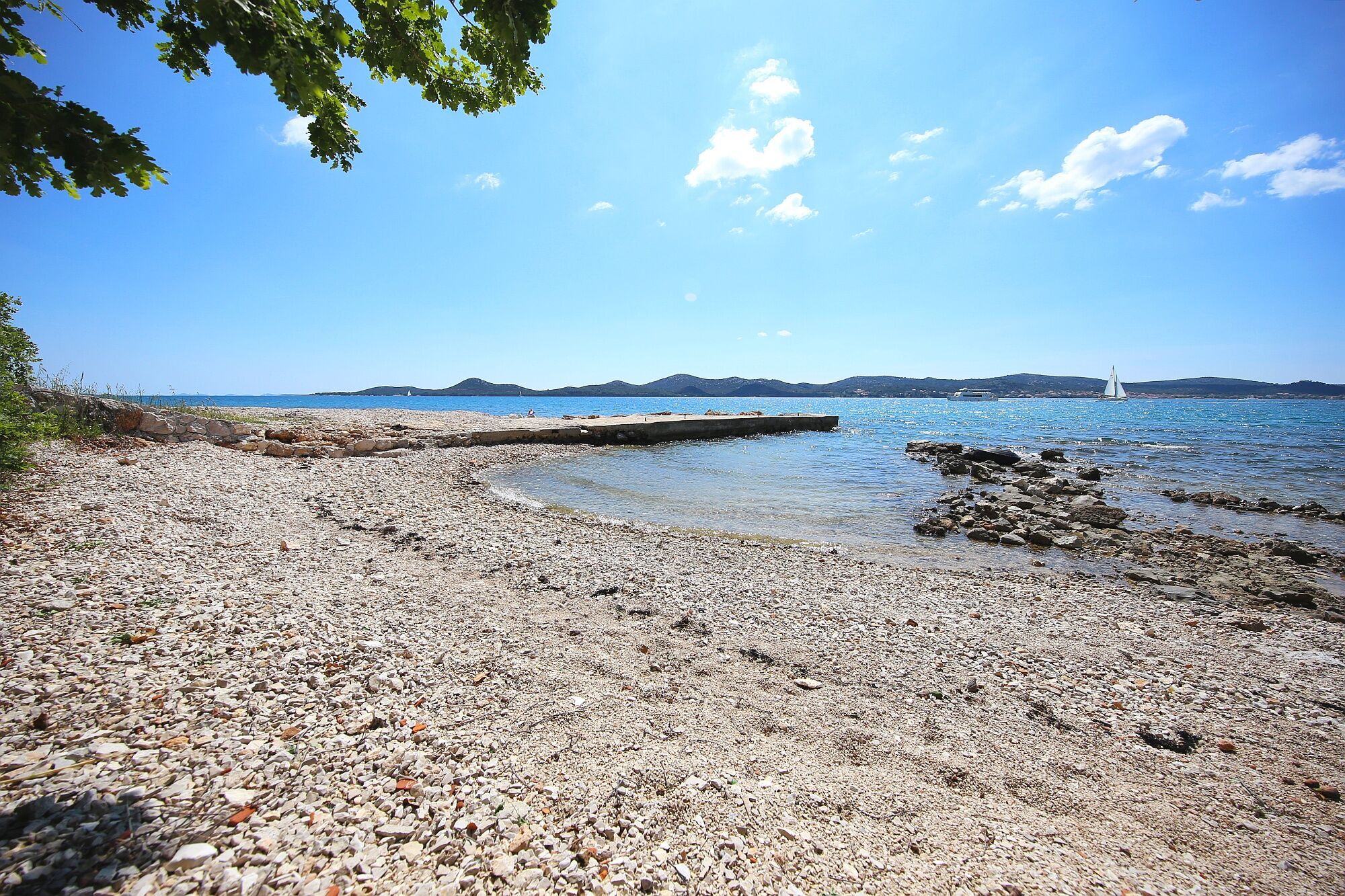 A pebbled beach with a pier extending into calm waters, surrounded by clear skies and distant hills.