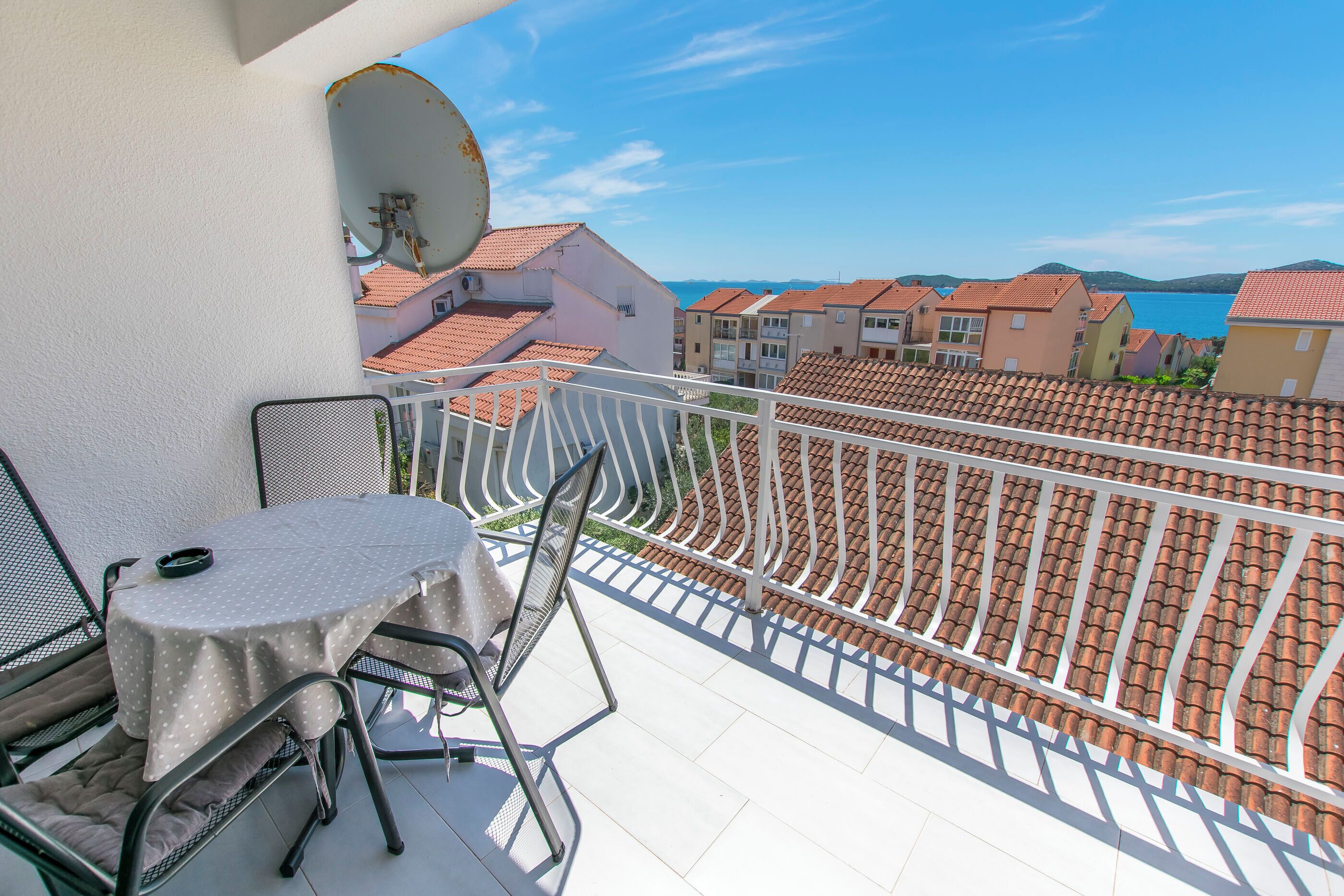 Un balcon avec une table et des chaises, une antenne parabolique, et une vue sur des bâtiments aux toits de tuiles et sur une étendue d’eau.