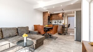 A modern living room with a sofa, ottoman, and coffee table. There is a dining area with chairs and a table. The kitchen is visible in the background with cabinets and a countertop. - Apartments Andrea (Sibenik)