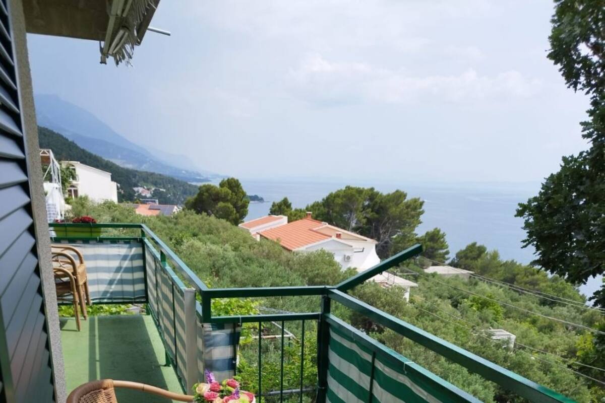 A balcony with a round table and wicker chairs, overlooking a scenic view of houses and trees.