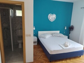 Ein Schlafzimmer mit einem Bett, einem weißen Nachttisch, einem an der Wand montierten Heizgerät und einem durch eine offene Tür einsehbaren Badezimmer.