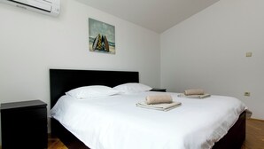 Una cama bien hecha con sábanas blancas y dos toallas, un mesita de noche negro, un cuadro con tablas de surf y un equipo de aire acondicionado en la pared.