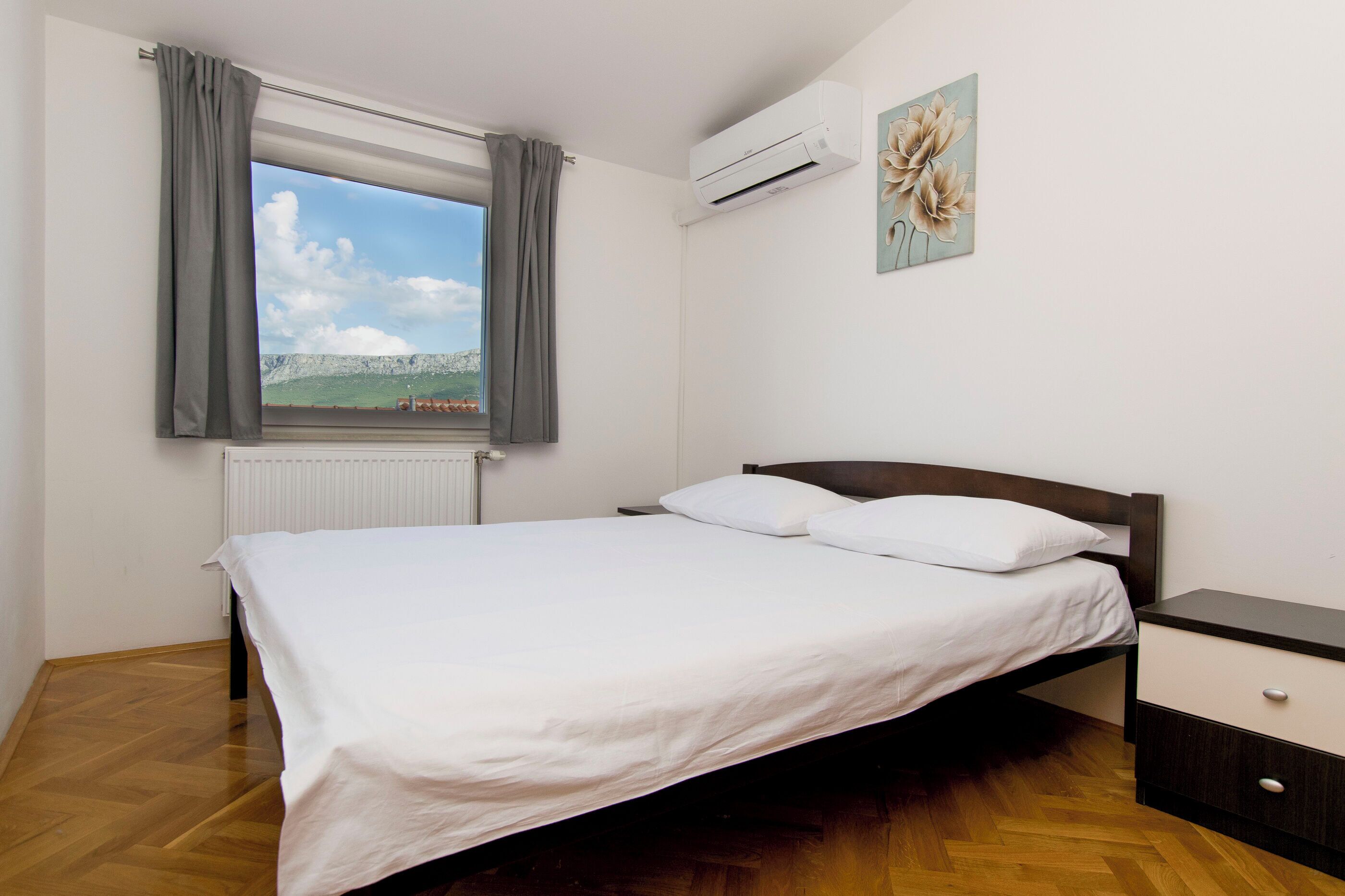A bedroom with a bed, a nightstand, a window with curtains, an air conditioner, and a wall-mounted picture.
