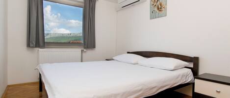 A bedroom with a bed, a nightstand, a window with curtains, an air conditioner, and a wall-mounted picture.