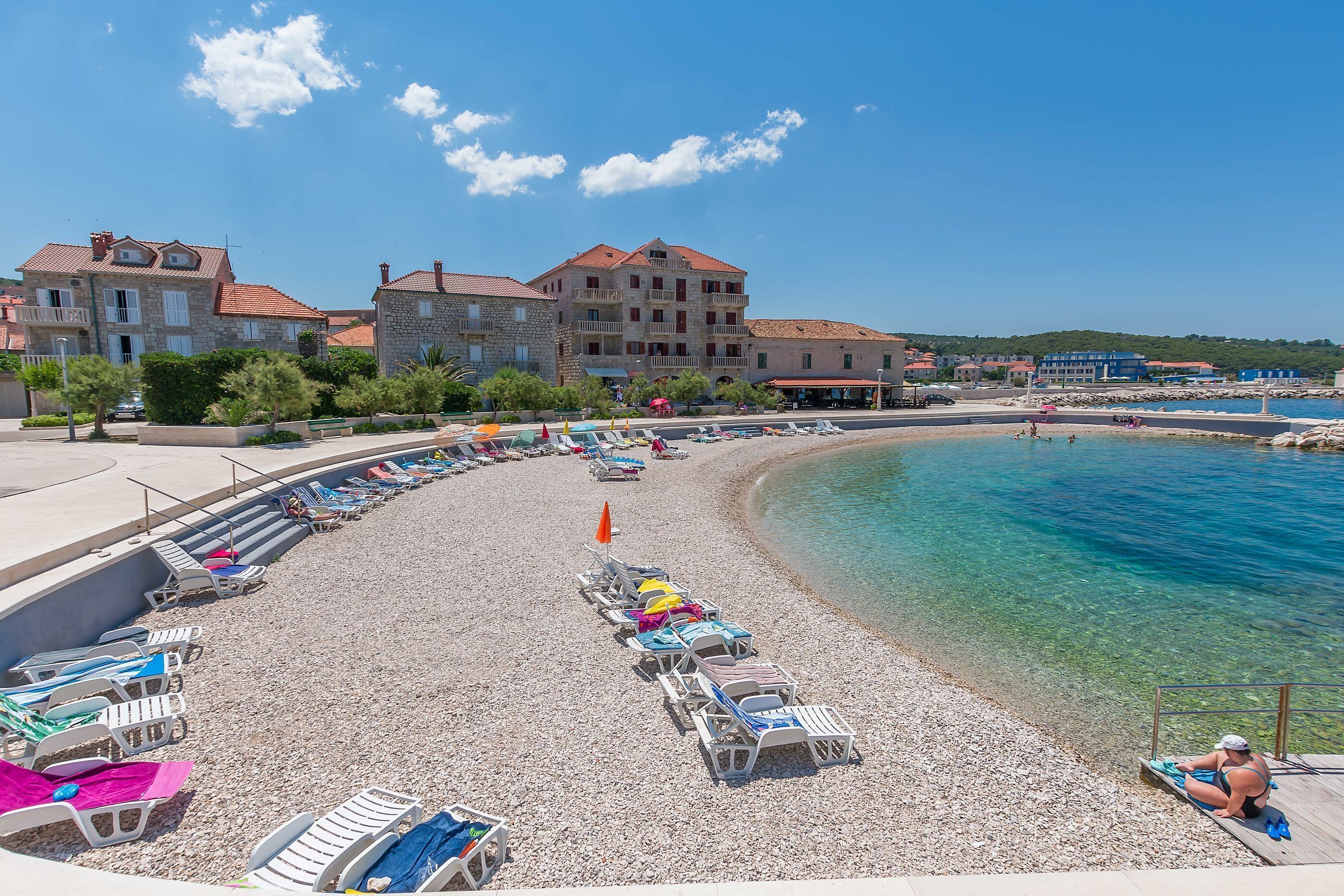 Ein Kiesstrand mit Liegestühlen und Sonnenschirmen, ein klares, blaues Schwimmbecken und Gebäude im Hintergrund.