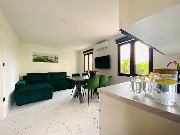 Una sala de estar moderna con un sofá verde, una mesa de comedor y sillas. Hay un televisor montado en la pared, un ventanal con vista a vegetación, y una zona de cocina con una encimera y vasos de agua.