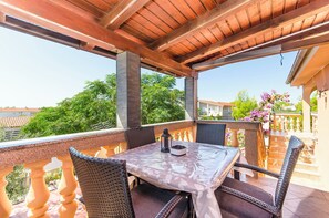 En overdækket terrasse med et bord og stole, en lygte og udsigt over det omkringliggende kvarter.