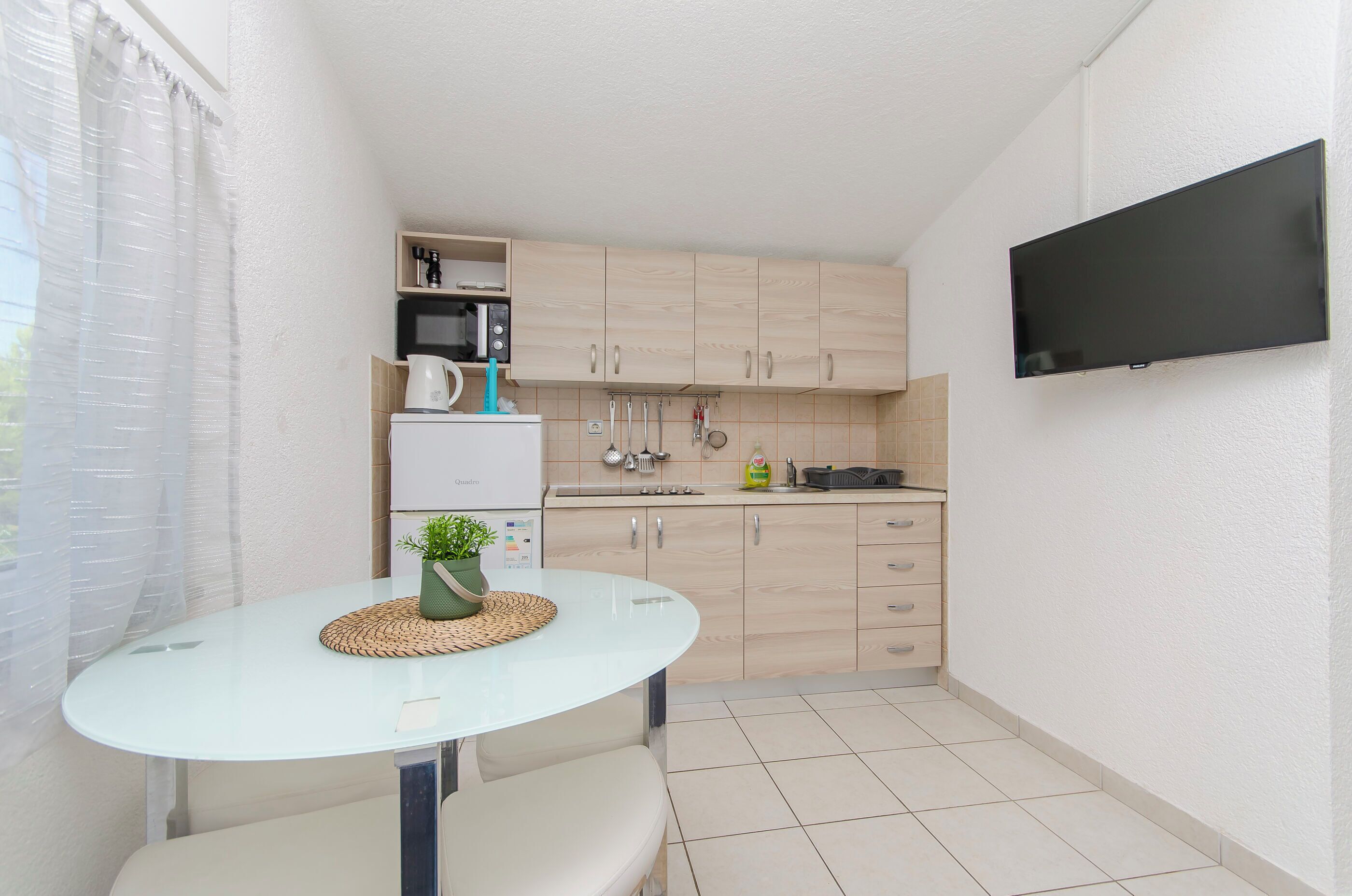 Ett kompakt kök med mikrovågsugn, vattenkokare och en platt-TV som är monterad på väggen.