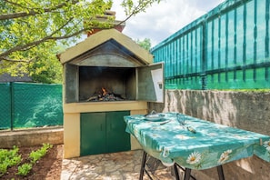 An outdoor barbecue area with a grill, a table with a floral tablecloth, and a green fence. - House Tamy - With Jacuzzi (Omis)