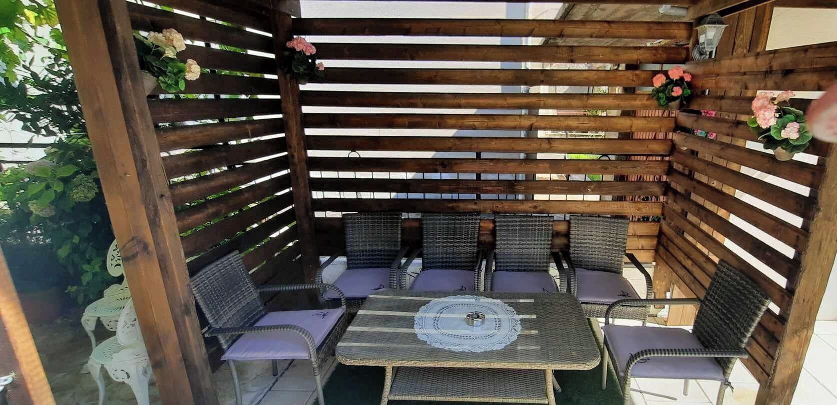 A wooden pergola with wicker furniture and cushions, surrounded by potted plants and flowers.