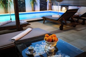 Un espace aménagé au bord de la piscine, avec des chaises longues, une table sur laquelle se trouve un saladier rempli d’oranges, et une table en verre avec deux tasses et une théière.