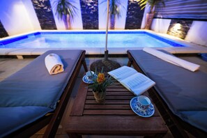 Een terras bij het zwembad met twee ligbedden, een tafel met een boek en een kopje, en een kleine potplant.