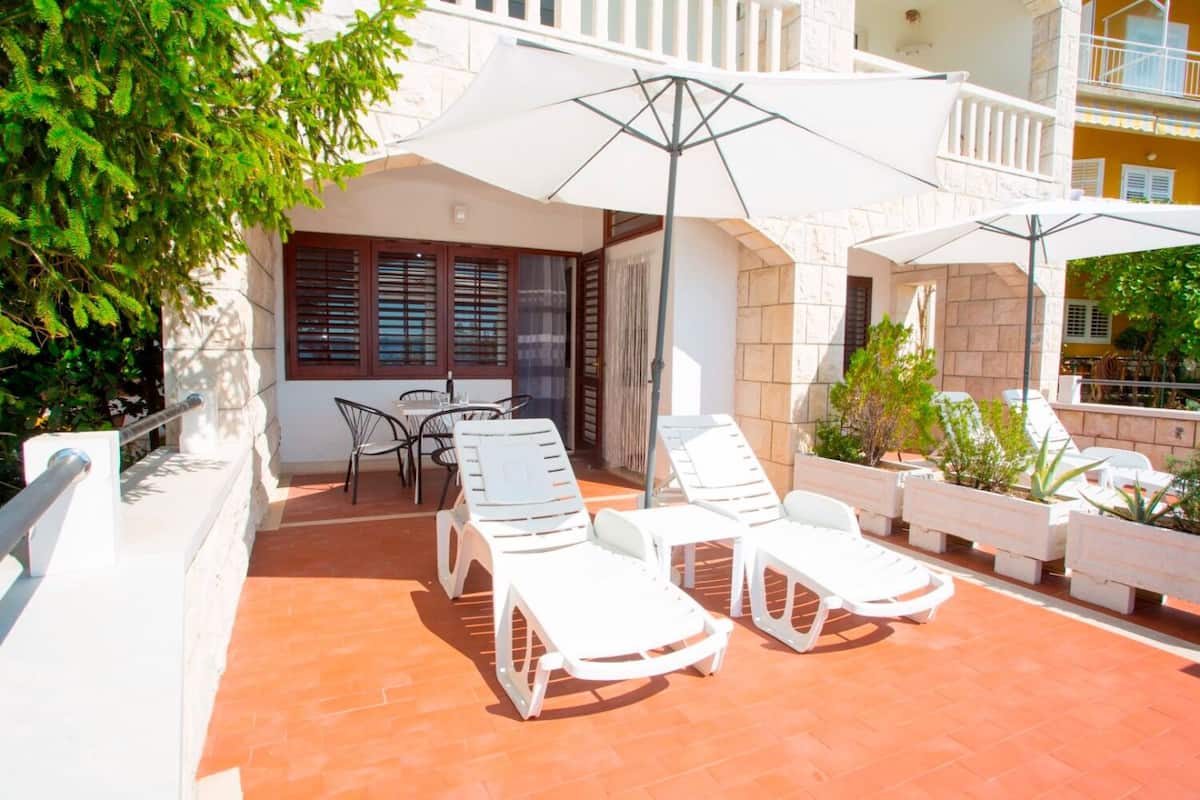 A terrace with white lounge chairs and a table, surrounded by potted plants and a white umbrella.