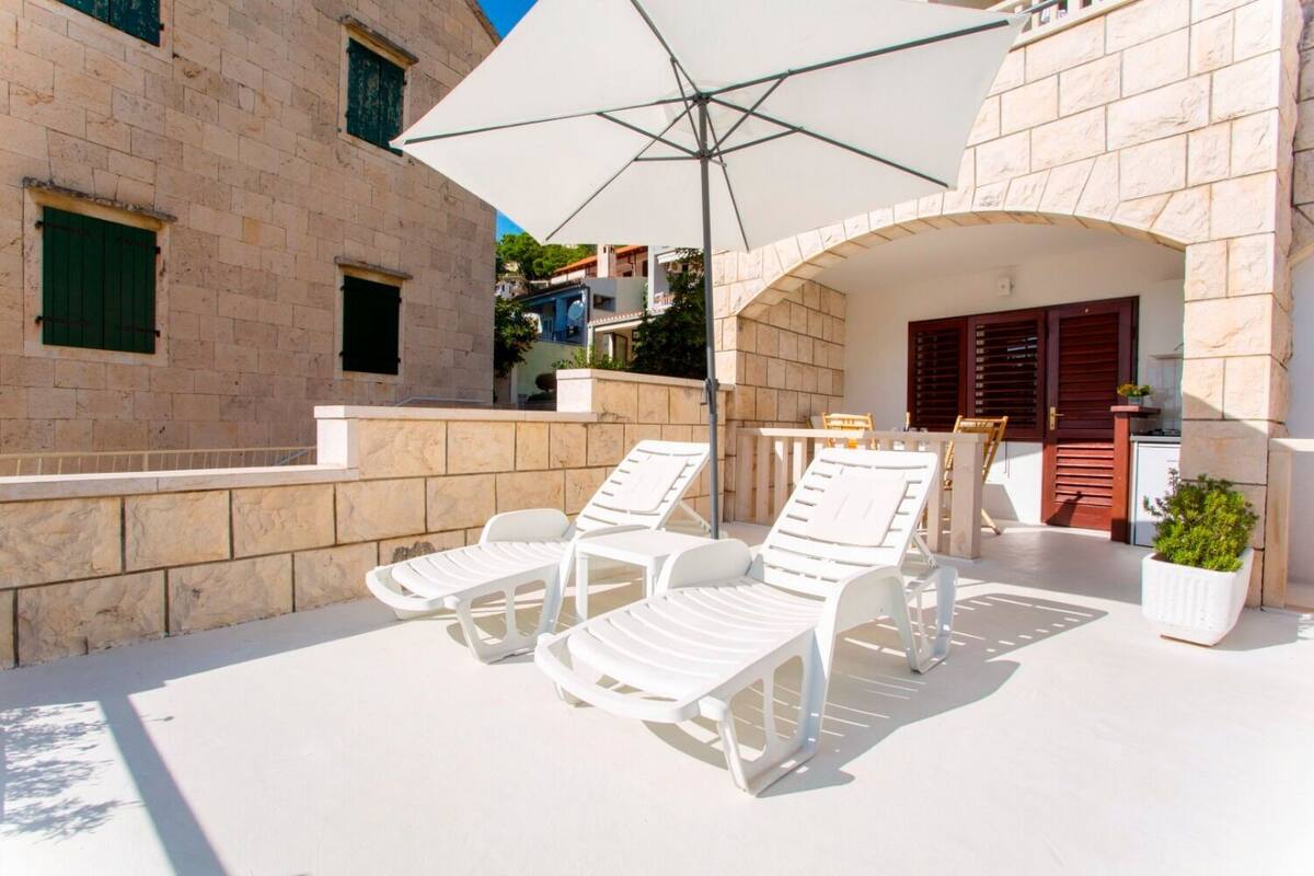 A sunlit outdoor seating area with white lounge chairs and a table, a stone building with a balcony, and a potted plant.