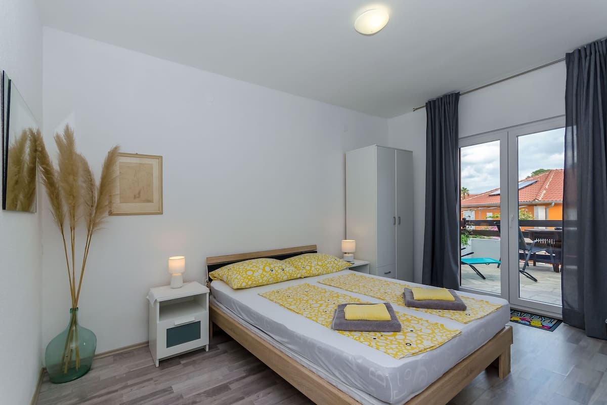 A bedroom with a bed, bedside tables, a nightstand, a vase with pampas grass, and a view of the outside.