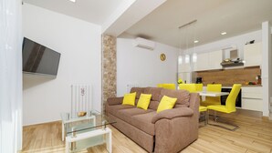 Een moderne woonkamer met een bruine bank, een glazen salontafel en gele stoelen. Een flatscreen televisie is aan de muur bevestigd, en er is een keuken zichtbaar op de achtergrond.