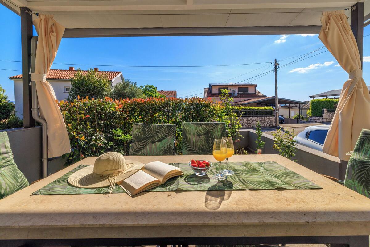A covered outdoor seating area with a table set for two, featuring a book, a hat, a bowl of strawberries, and a glass of orange juice.