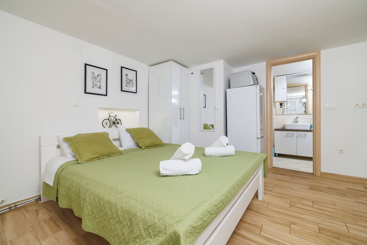 A bedroom with a bed, green bedspread, white pillows, and two rolled towels. There is a white wardrobe, a mirror, and a white cabinet with a sink.