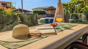Een zonnig buitenzitgedeelte met een tafel, een hoed, een bril, een boek, een glas sinaasappelsap en een schaal aardbeien.