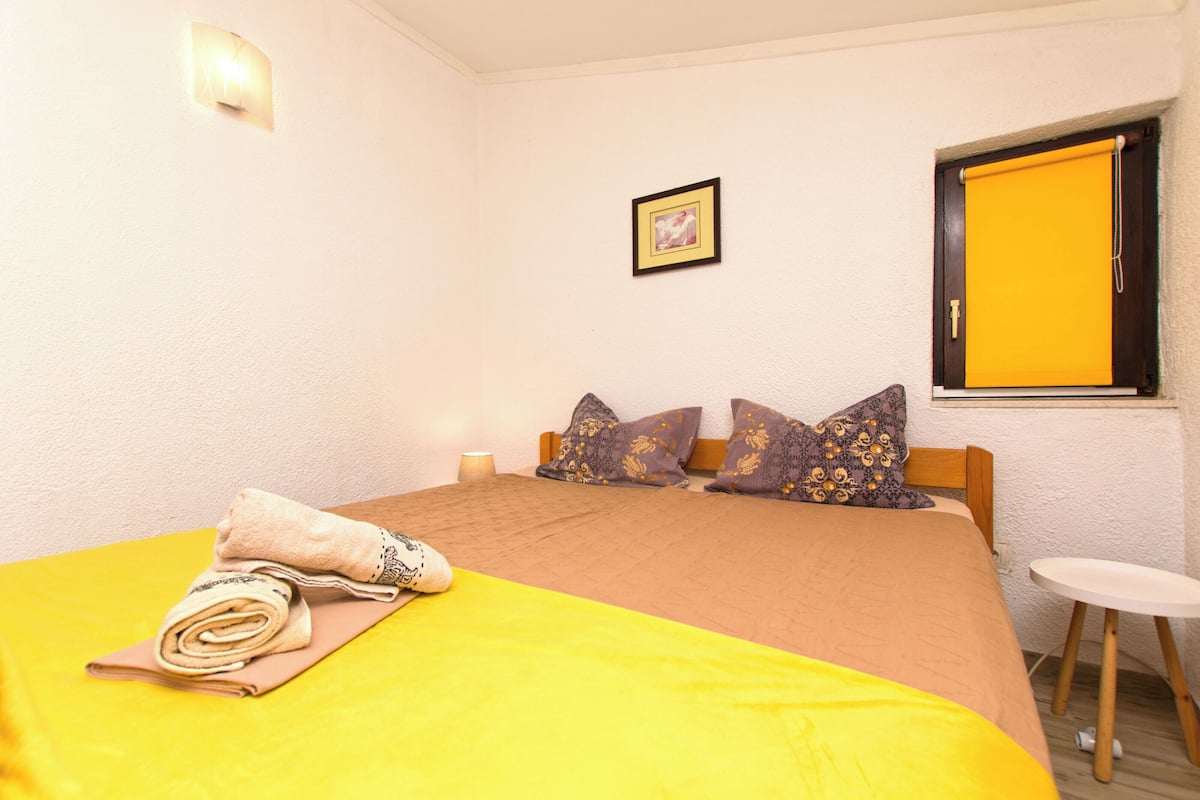A bedroom with a bed, pillows, a bedside table, and a window with a yellow blind.