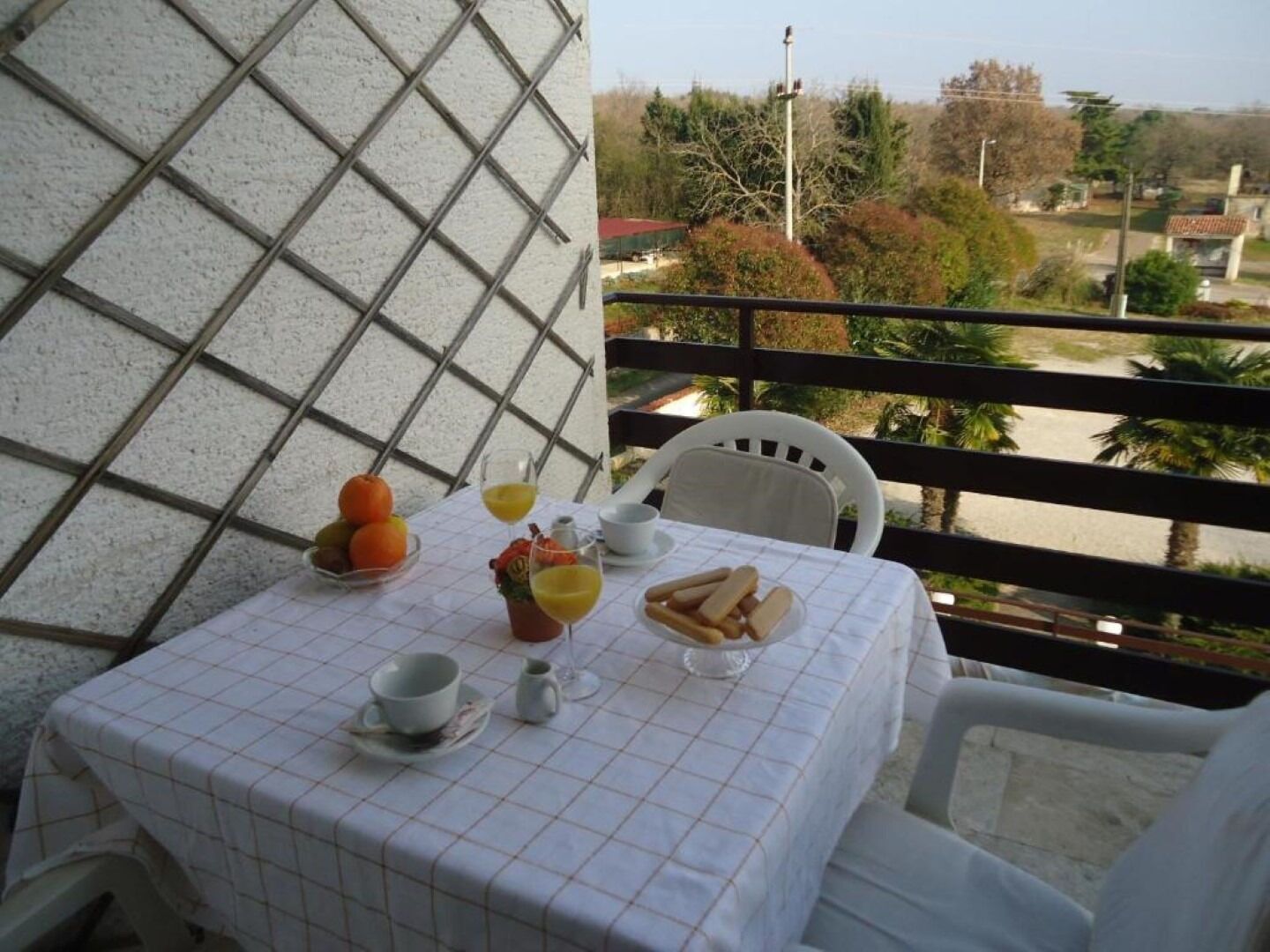 Une table dressée pour le petit-déjeuner, avec des oranges, du pain et des boissons, sur un balcon donnant sur un quartier résidentiel.