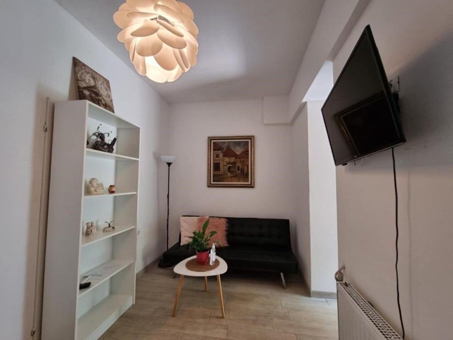 A living room with a black sofa, a white shelving unit, a round table with a plant, a painting on the wall, and a ceiling light fixture.
