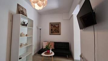 A living room with a black sofa, a white shelving unit, a round table with a plant, a painting on the wall, and a ceiling light fixture.