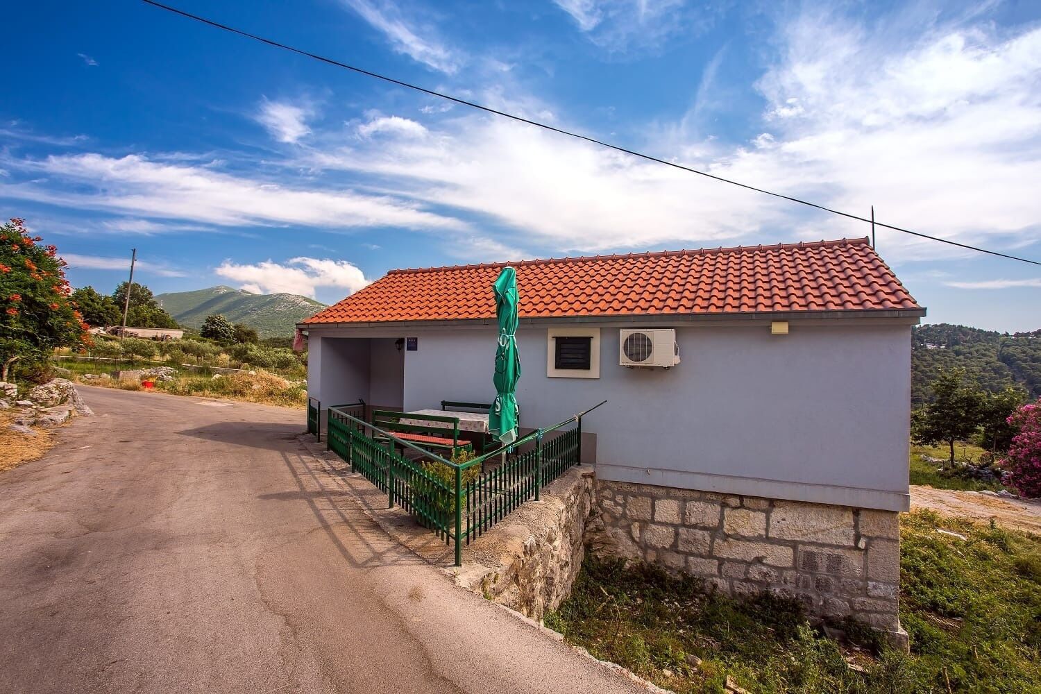 A small, single-story building with a red-tiled roof and a stone foundation. It features a green outdoor seating area with a table and chairs, and an air conditioning unit on the wall.