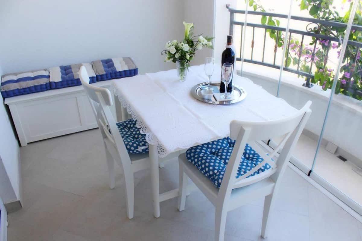Un coin repas avec une table et des chaises blanches, un plateau avec une bouteille et un verre, et un banc agrémenté d’un coussin bleu et blanc.