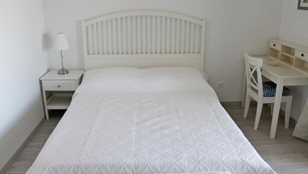 Una cama blanca con cabecera, una mesita de noche con lámpara y un pequeño escritorio con silla.