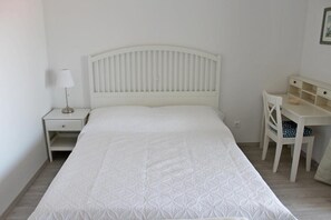 Una cama blanca con cabecera, una mesita de noche con lámpara y un pequeño escritorio con silla.