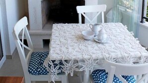 Un comedor con una mesa blanca dispuesta para cuatro personas, sillas blancas con cojines azules, una repisa de chimenea blanca con un reloj y vistas a través de una ventana con cortinas translúcidas.