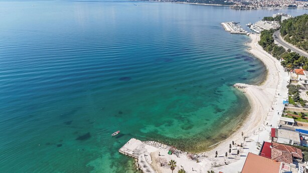 An aerial view of a coastal area with a clear beach, a marina with boats, and buildings along the shore.