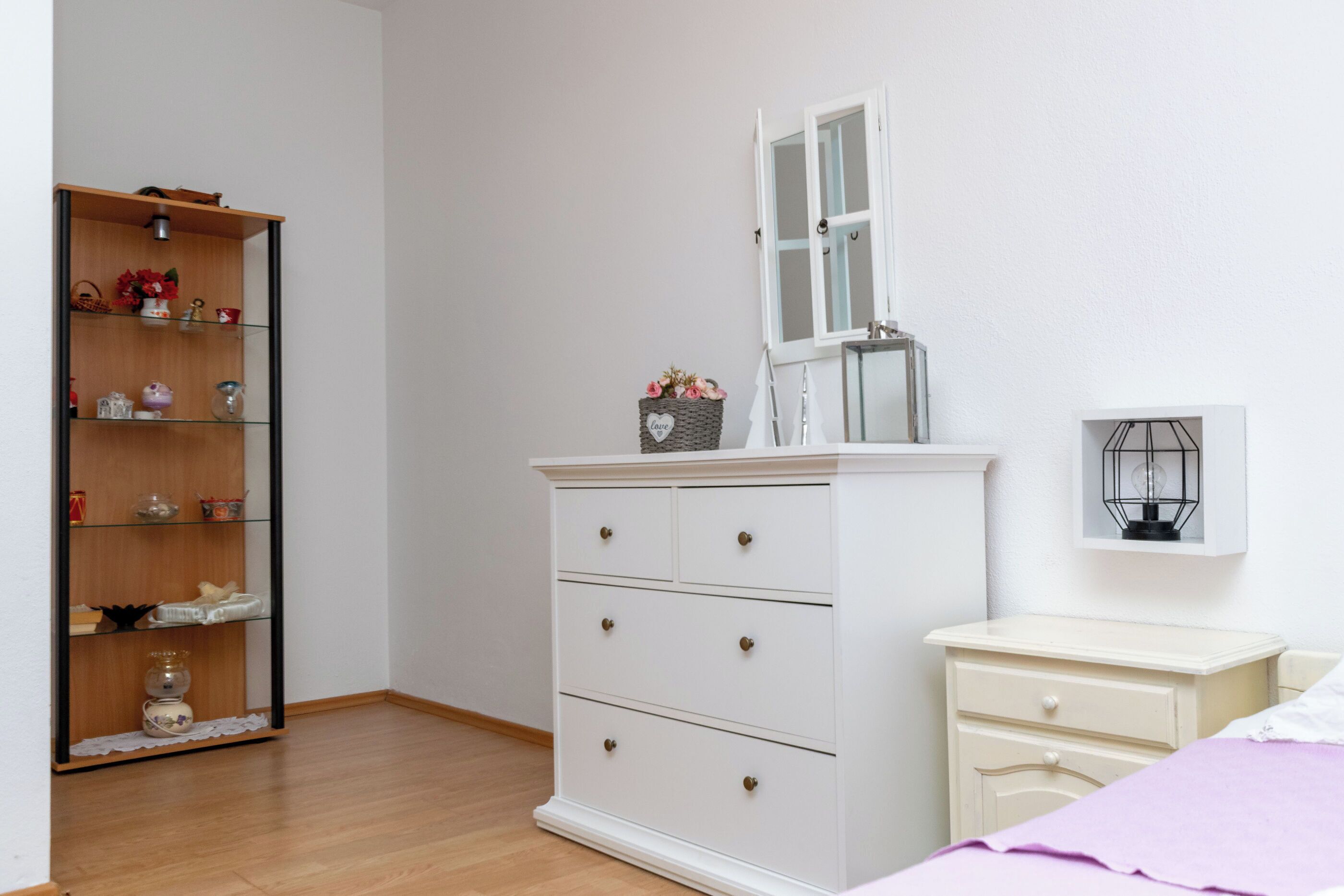 A bedroom with a white dresser, a nightstand, a bed with a purple blanket, a wooden floor, a white wall, and a window with a white frame.