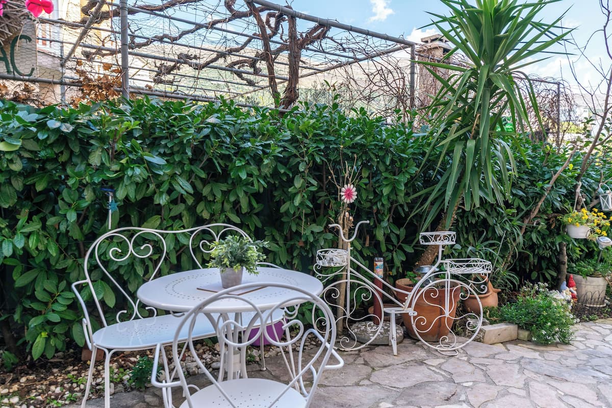 A patio with a metal table and chairs, surrounded by lush greenery and a trellis structure.