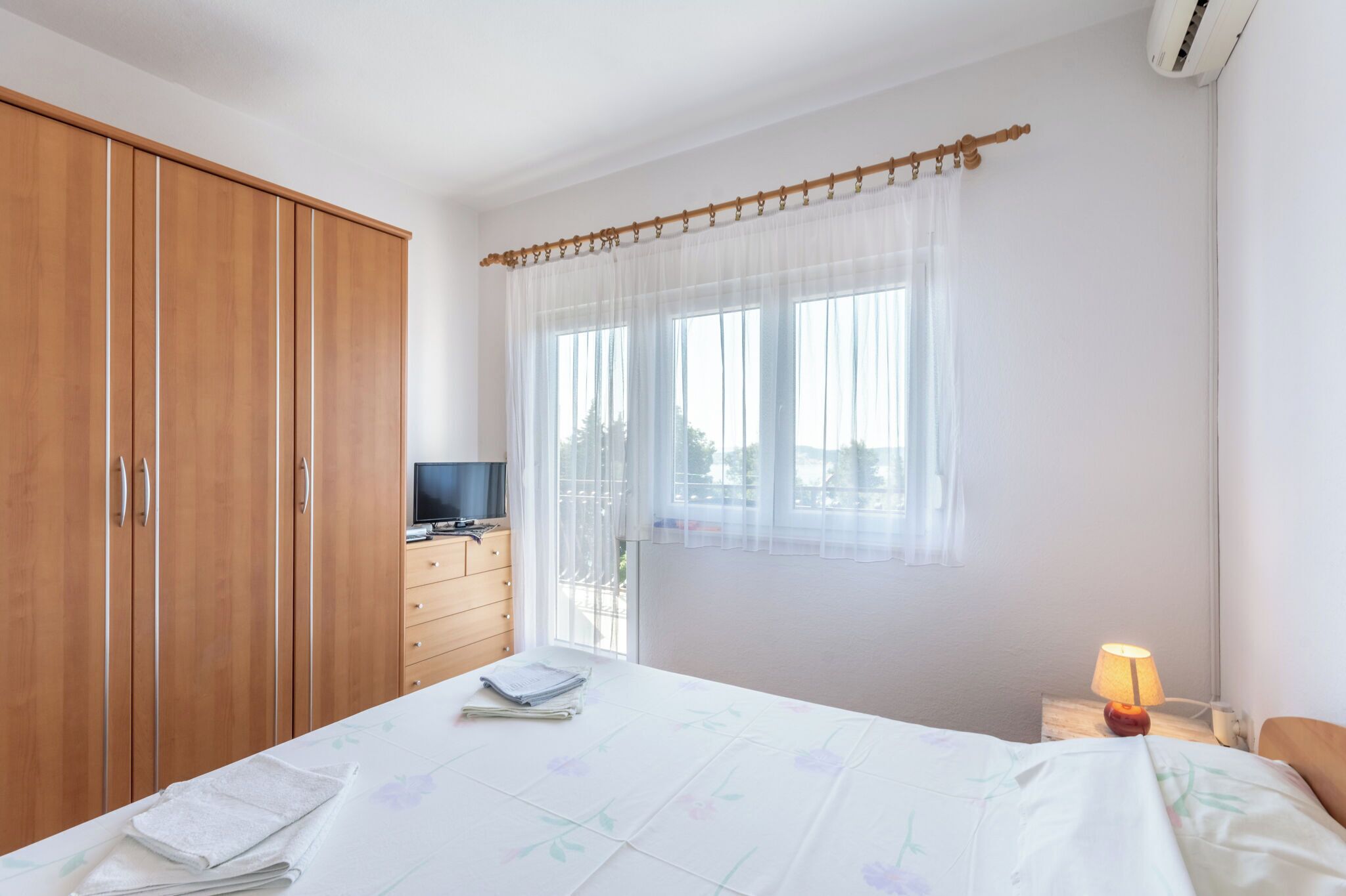 A bedroom with a bed, a wooden wardrobe, a desk with a lamp, and a window with sheer curtains.