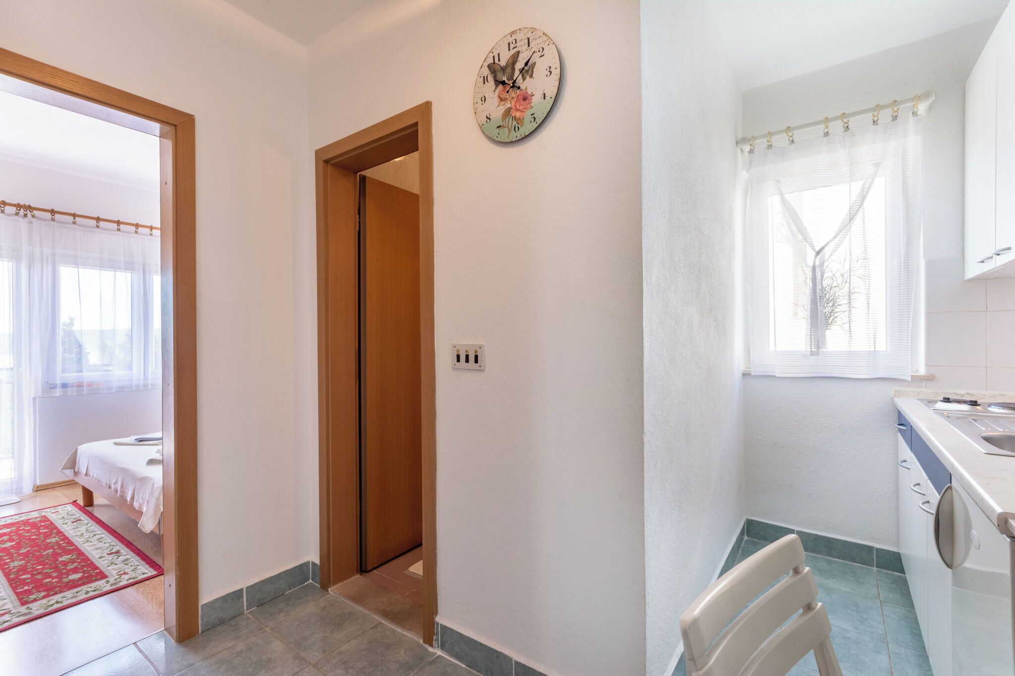 Habitació amb un llit, una porta de fusta, un rellotge de paret, una finestra amb cortina i una cuina amb una pica i armaris.