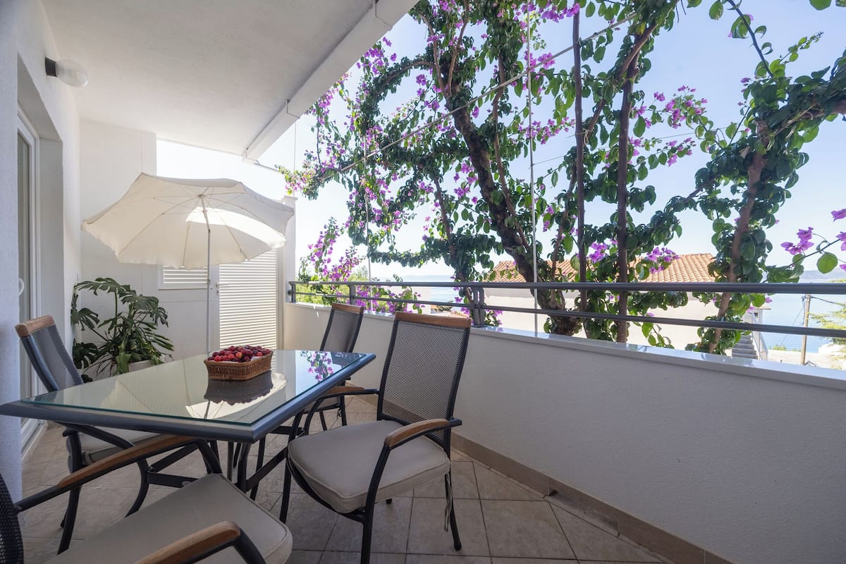 A balcony with a glass table and chairs, a potted plant, and a view of the sea.