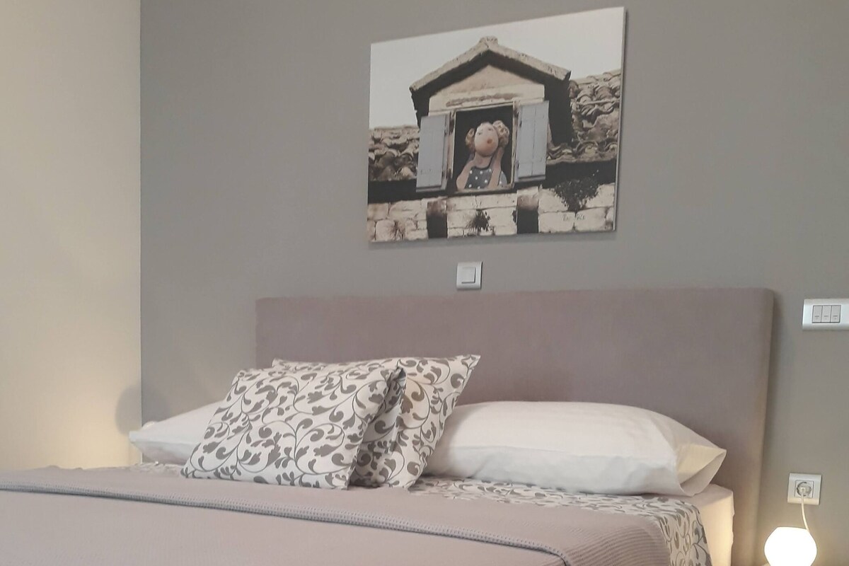 Ein ordentlich bezogenes Bett mit einem gemusterten und einem weißen Kissen, einer Nachttischlampe sowie einem Wandbild.