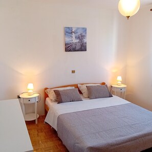 A bedroom with a bed, two bedside tables with lamps, a nightstand, a wall-mounted painting, and a wooden floor. - Apartment Sunset Lina (Pula)