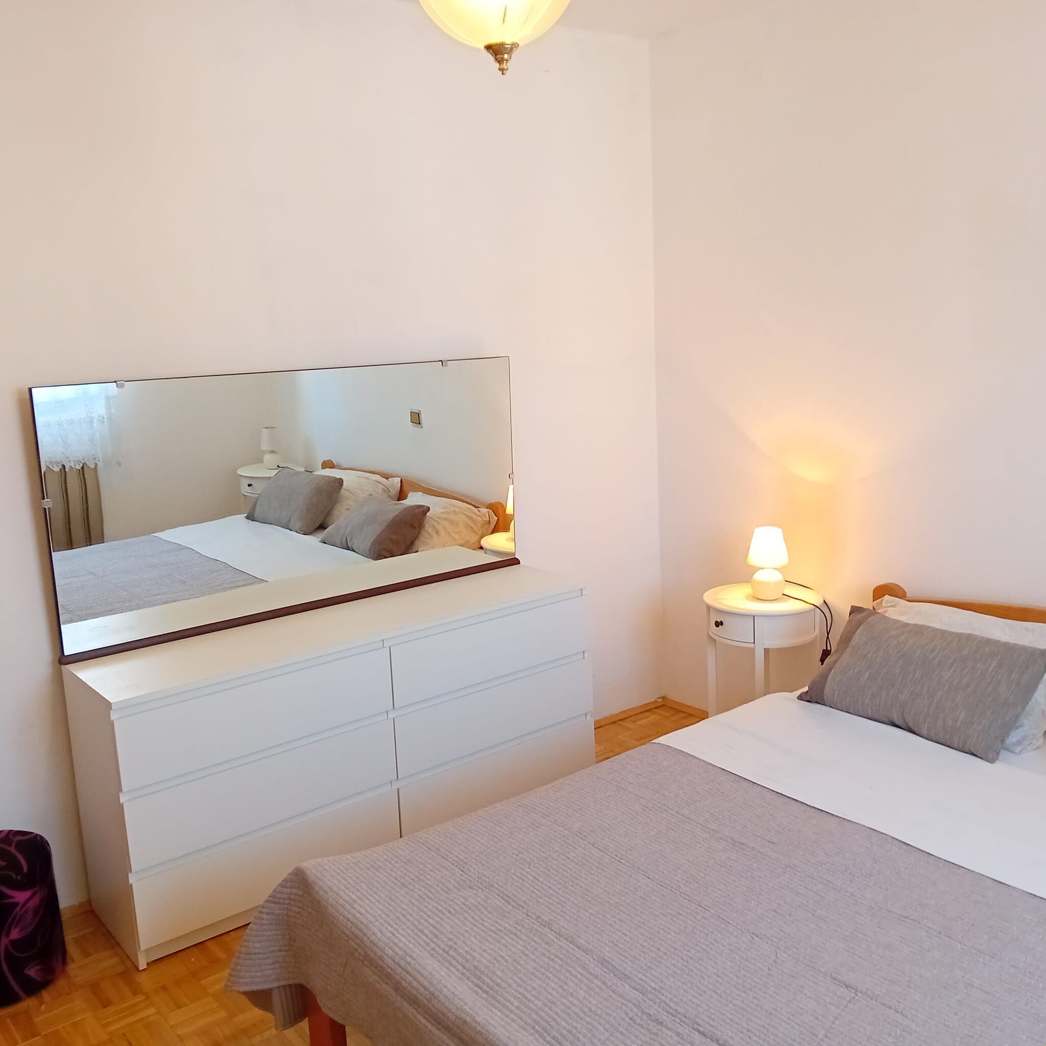 A bedroom with a large mirror, a bedside table with a lamp, a dresser, and a bed with pillows.