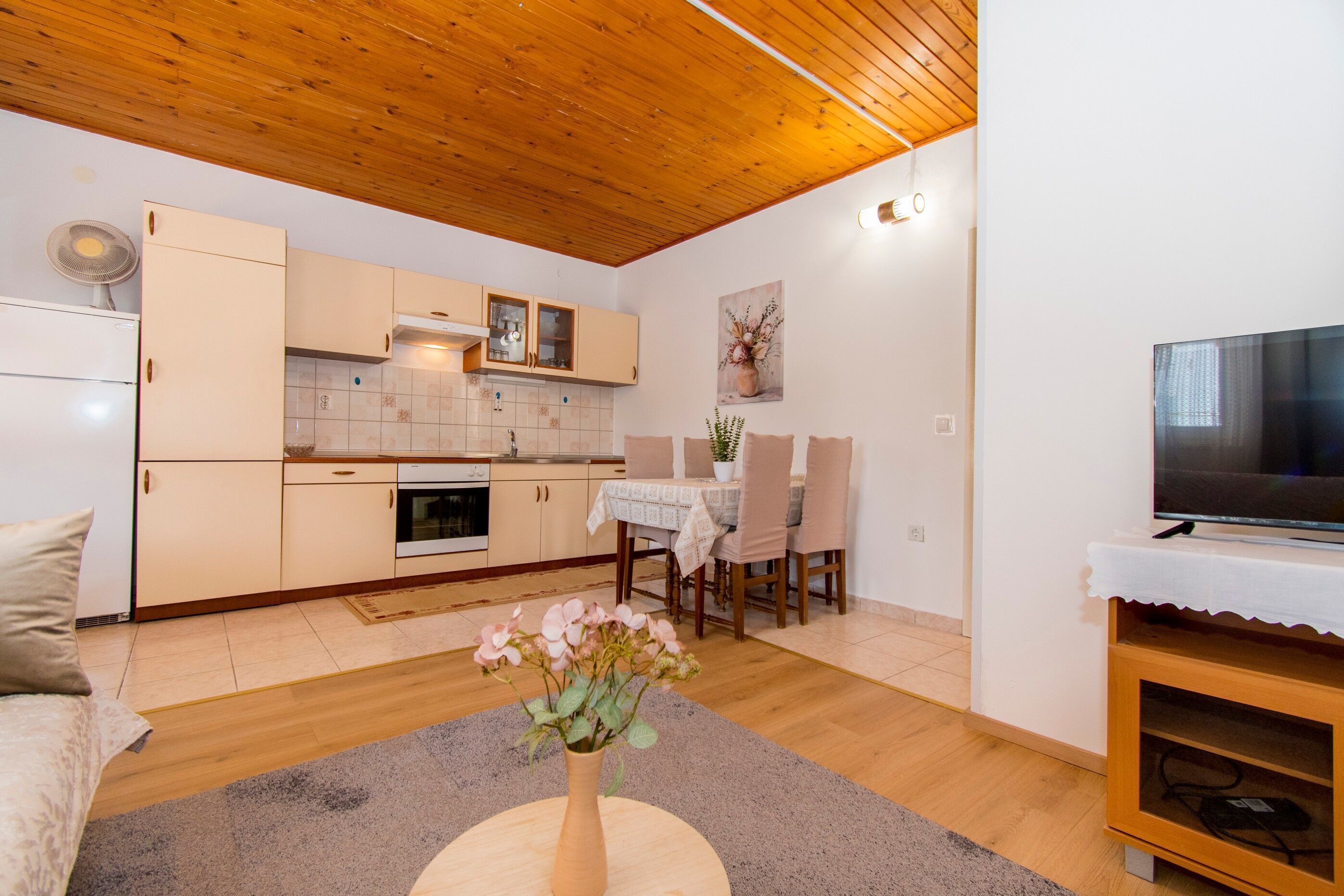 A modern kitchen with wooden cabinets, a dining table with chairs, a TV on a stand, and a vase with flowers on the floor.