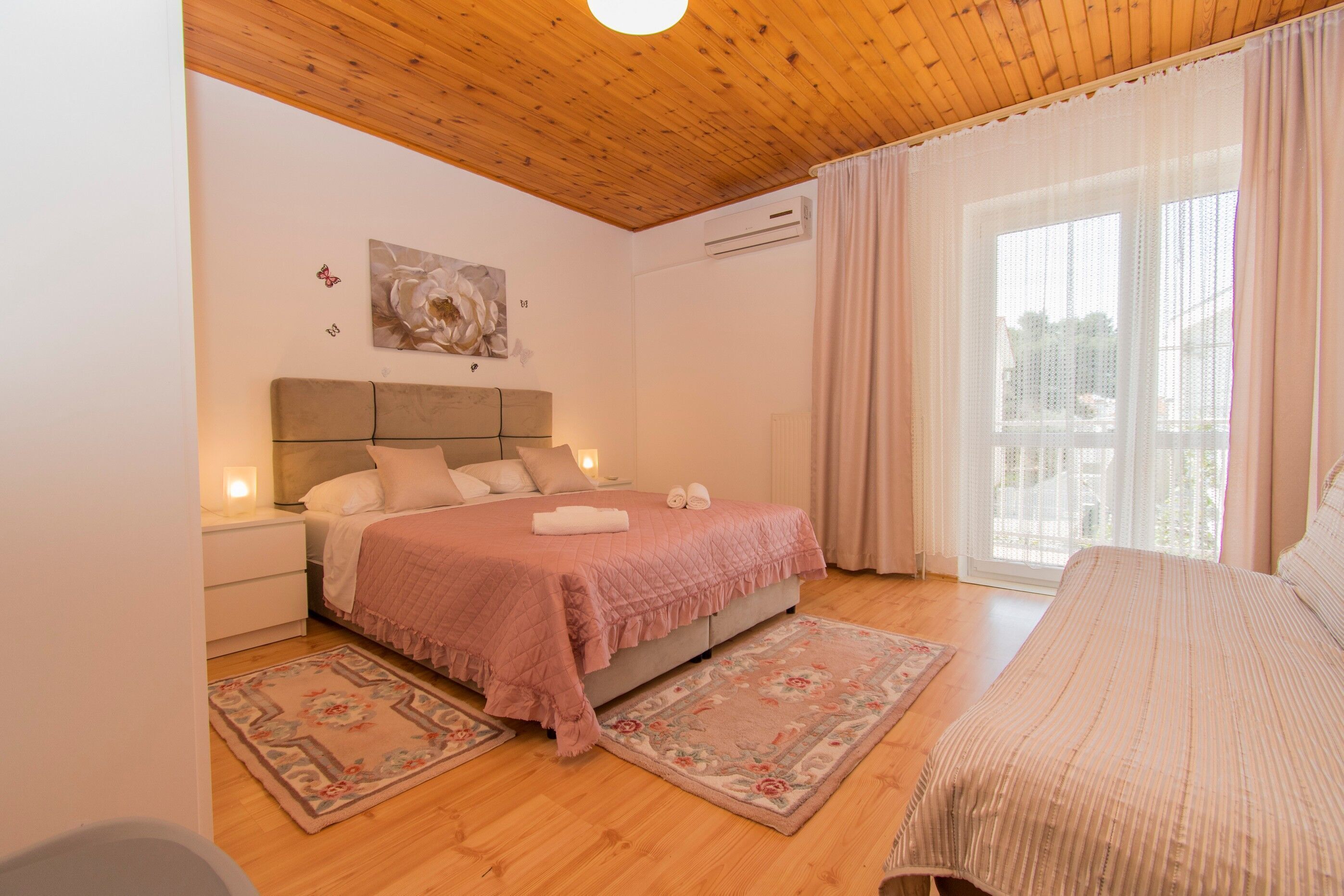 A bedroom with a wooden ceiling, a bed with a pink quilt, two nightstands, a wall-mounted air conditioner, and a large window with curtains.