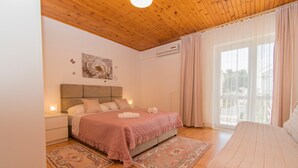 A bedroom with a wooden ceiling, a bed with a pink quilt, two nightstands, a wall-mounted air conditioner, and a large window with curtains. - Apartments Neve (Vodice)
