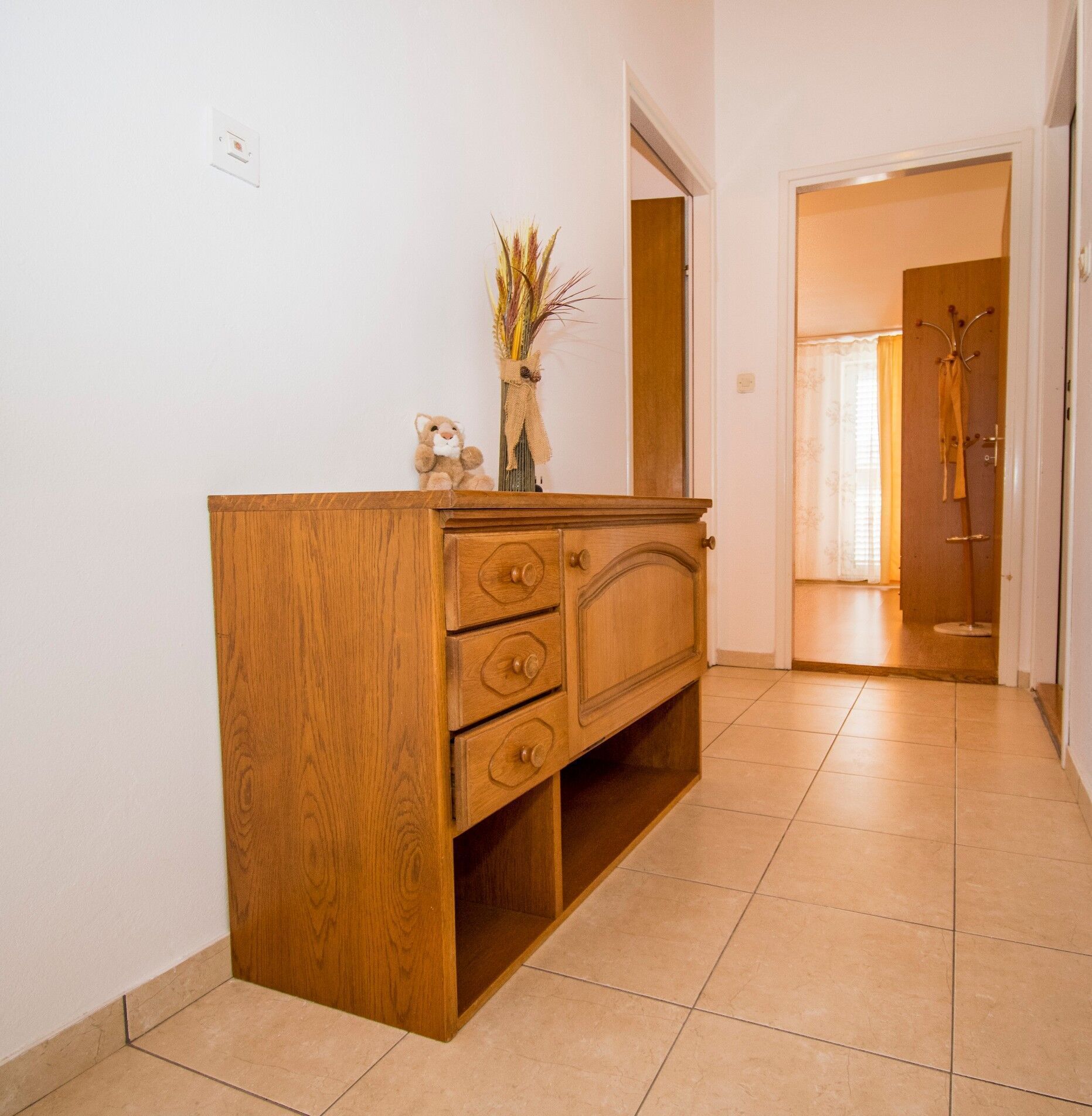A wooden dresser with three drawers and a mirror above it, placed against a white wall in a hallway with tiled flooring.